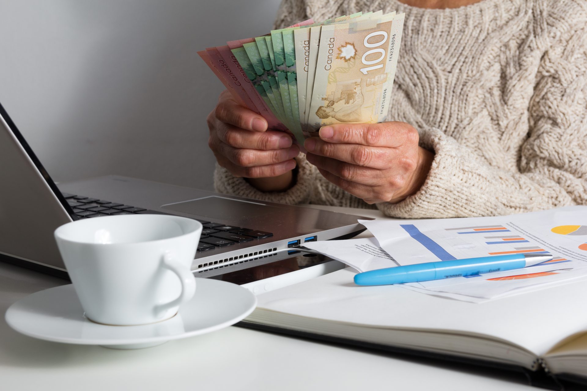 Woman counting Canadian money beside a laptop, coffee mug, and spreadsheets